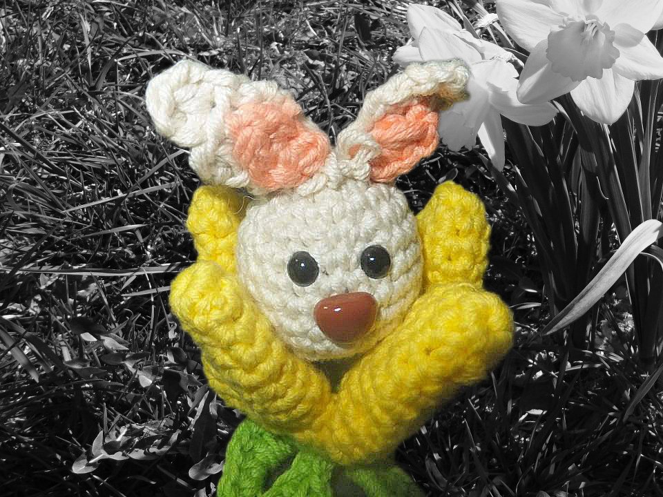 Gehäkelter Osterhase mit gelben Armen, weißem Kopf und pfirsichfarbenen Ohren vor monochromem Gras und Narzissen