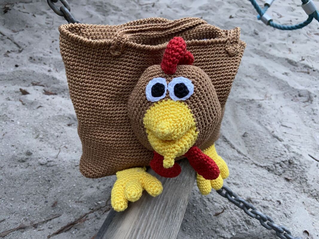 Verspielte gehäkelte Tasche mit großem Amigurumi-Hühnerkopf, gelbem Schnabel, rotem Kamm und gelben Füßen auf sandigem Boden
