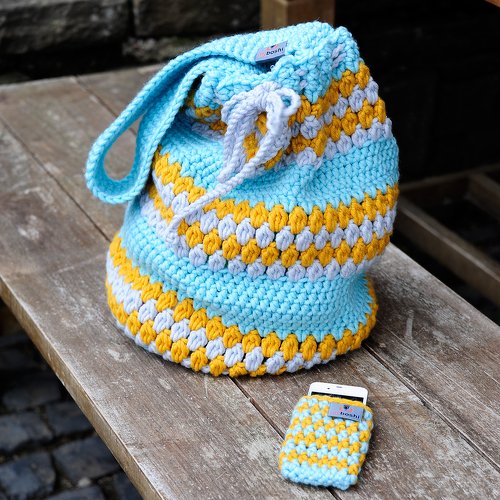 Gehäkelte Zugbeuteltasche und Handyhülle in Hellblau, Senfgelb und Hellgrau auf Holztisch.