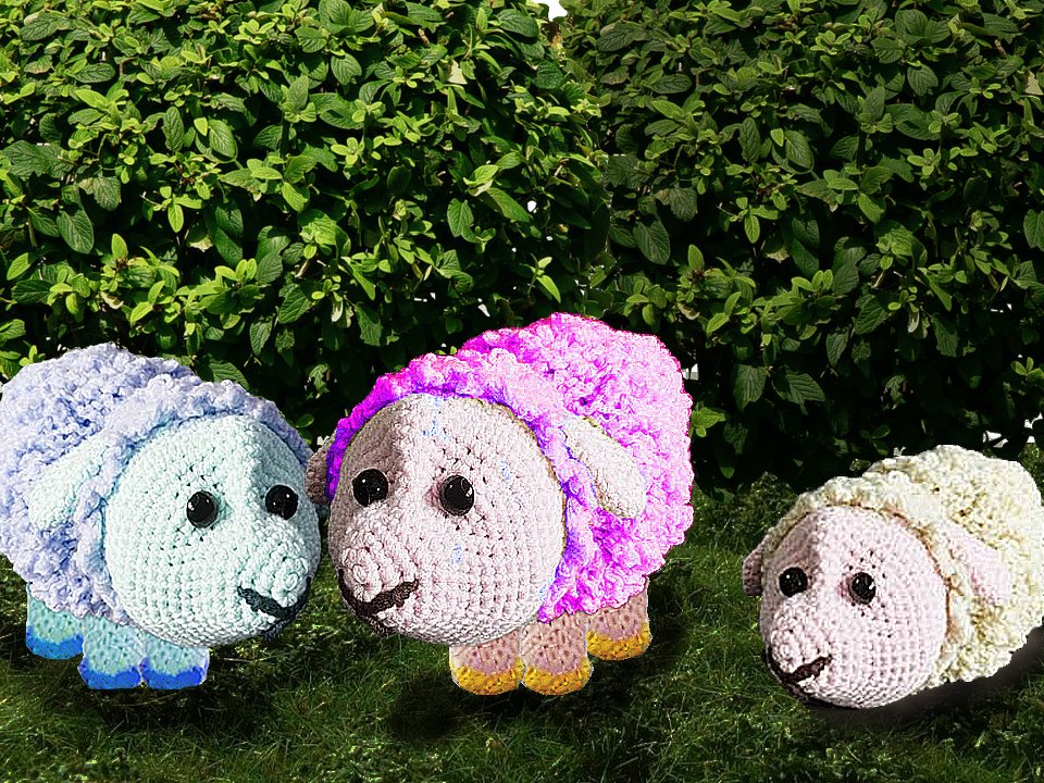 Drei Amigurumi-Schafe in Hellblau, Rosa und Cremeweiß auf Gras vor einem grünen Busch