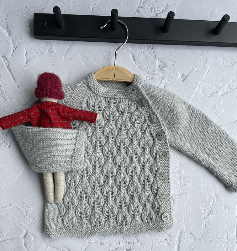 Gestrickter hellgrauer Babycardigan mit dekorativen Blattreihen, seitlicher Knopfleiste, Stoffpuppe in Tasche, aufgehängt an Holzbügel.