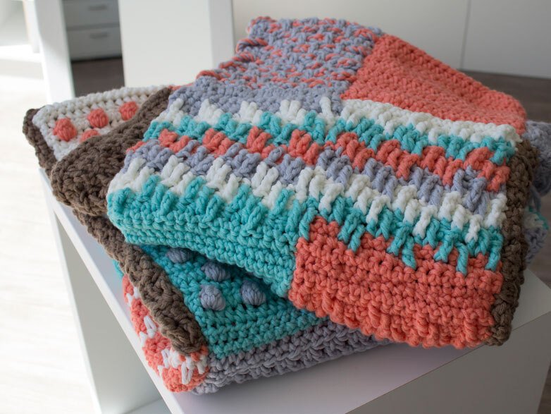 Gefaltete gehäkelte Decken mit Streifen- und Noppenmustern in Korall, Türkis, Weiß, Grau und Braun.
