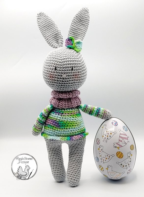 Grau gehäkelter Amigurumi-Hase mit buntem Pullover und Schleife, daneben ein Osterei mit Hühnermotiven.