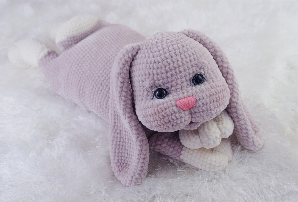 Lilac crocheted amigurumi rabbit lying on white fluffy fabric with long ears and pink nose