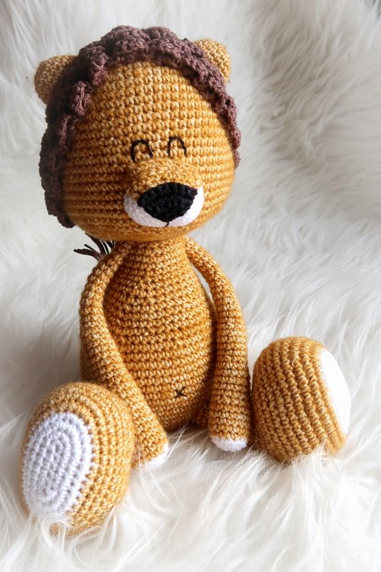 Sitzender Amigurumi-Löwe aus goldbraunem Garn mit brauner Mähne und weißen Fußsohlen auf weißem, flauschigem Untergrund.