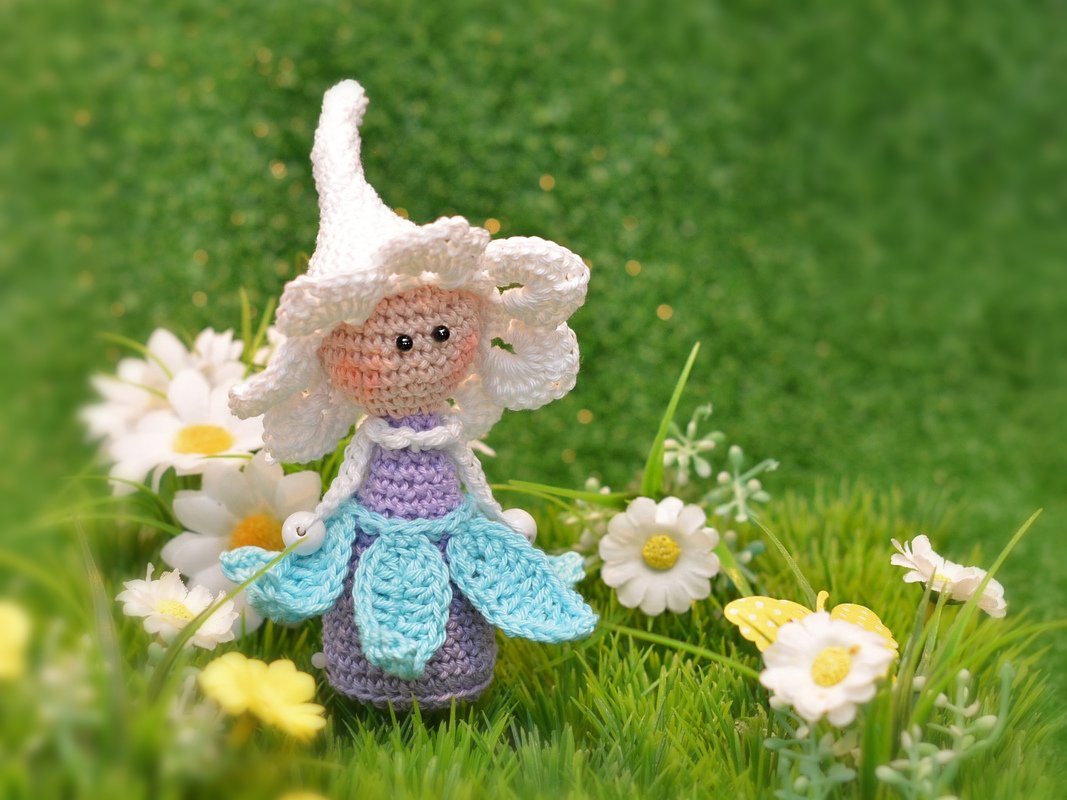 Gehäkelte Amigurumi-Blumenfee mit weißer Blütenmütze und hellblauem Blätterrock auf grüner Wiese mit Gänseblümchen.