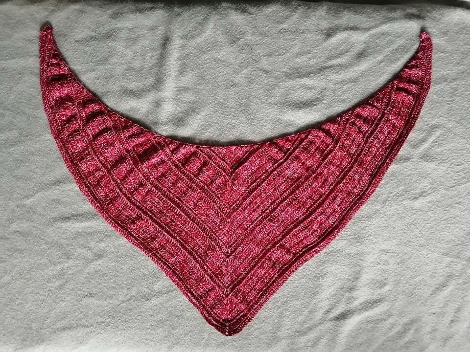 Handgehäkeltes Dreieckstuch in Rot‑Pink mit strukturierter Musterung auf hellgrauem Untergrund.