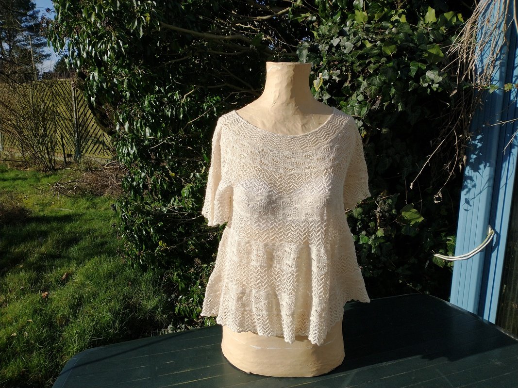 Cremefarbenes, gestricktes Damen‑Shirt mit kurzen Ärmeln und ausgestelltem Saum auf einer Schneiderpuppe vor Pflanzen.