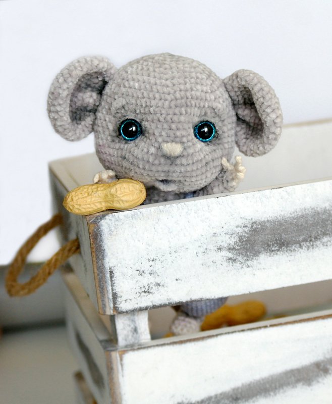 Grey crocheted amigurumi mouse with blue safety eyes holding a peanut in a whitewashed wooden crate
