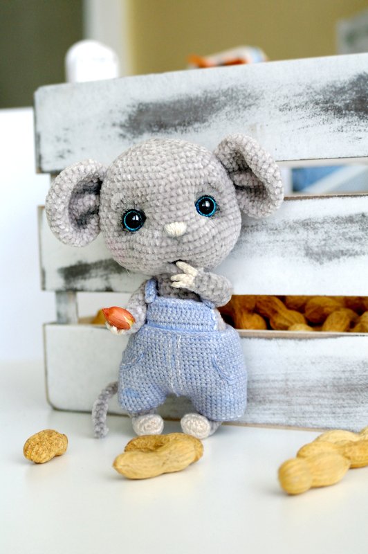 Gray crocheted amigurumi mouse wearing blue overalls, holding a peanut, standing in front of a wooden crate of peanuts