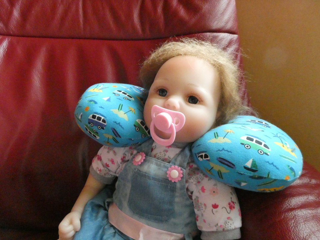 Puppe mit blauem U-förmigem Nackenkissen mit Wohnmobil- und Bootsmotiven auf rotem Sessel