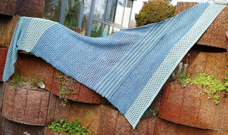 Gestricktes blau-minzgrünes Dreieckstuch mit Streifen-, Struktur- und Lochmustern auf einer Mauer.
