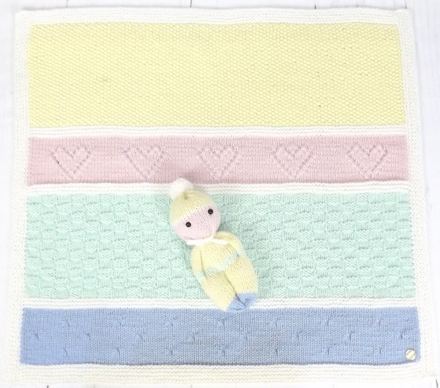 Draufsicht auf gestreifte Babydecke in Pastellfarben mit einer kleinen gestrickten Puppe; Details mit Herzmuster und Waffelstruktur sichtbar.