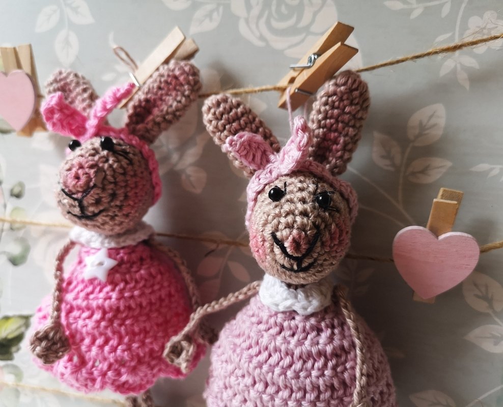 Zwei gehäkelte rosa Amigurumi-Hasen mit rosa Häkelmützen, aufgehängt an einer Schnur mit Wäscheklammern und Holzherzen
