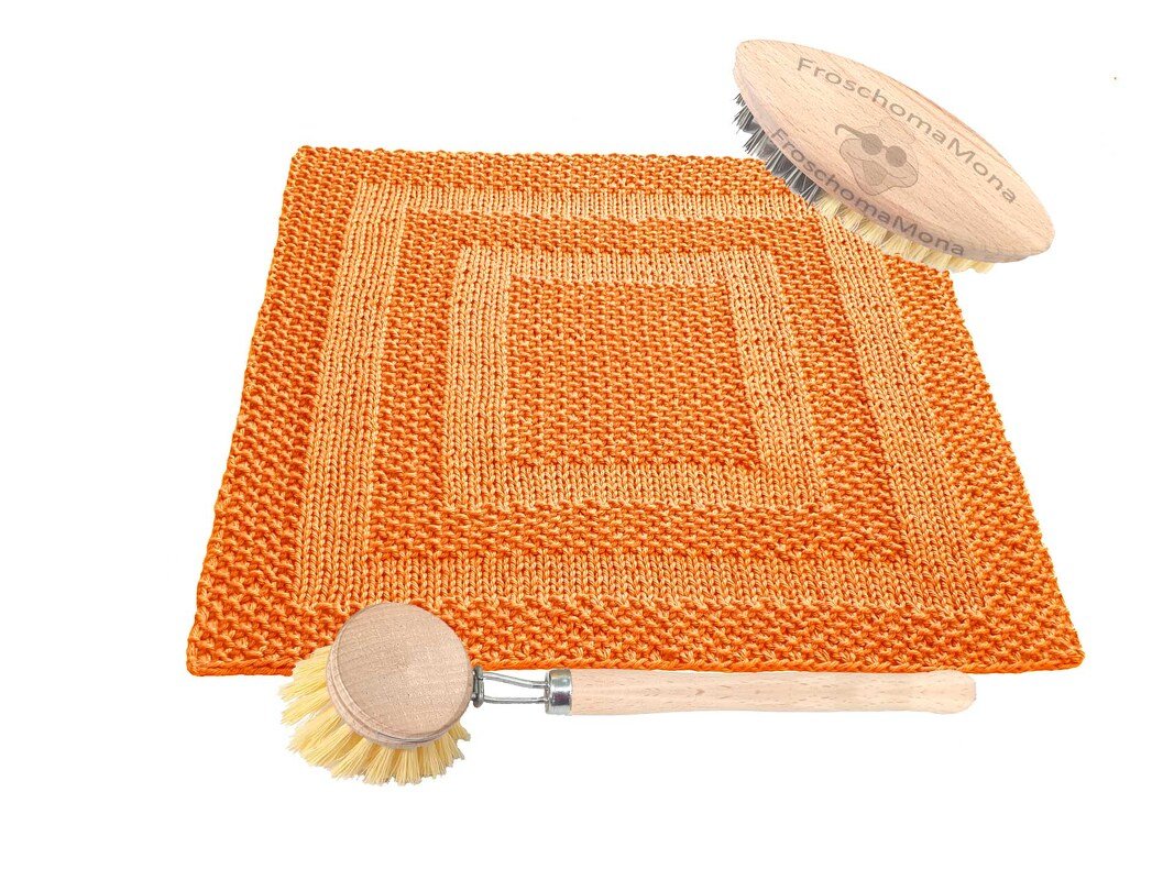 Quadratisches orange gestricktes Spültuch mit hellem Quadratmuster. Daneben liegen eine runde Spülbürste und eine längliche Reinigungsbürste aus Holz.