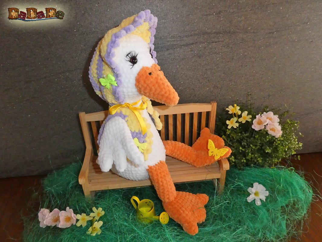 Handgearbeitetes Amigurumi: weiße Gans mit orangefarbenen Beinen und Schnabel sowie gestreifter gelb-lila Mütze, sitzend auf einer kleinen Holzbank neben künstlichen Blumen und einer Gießkanne.