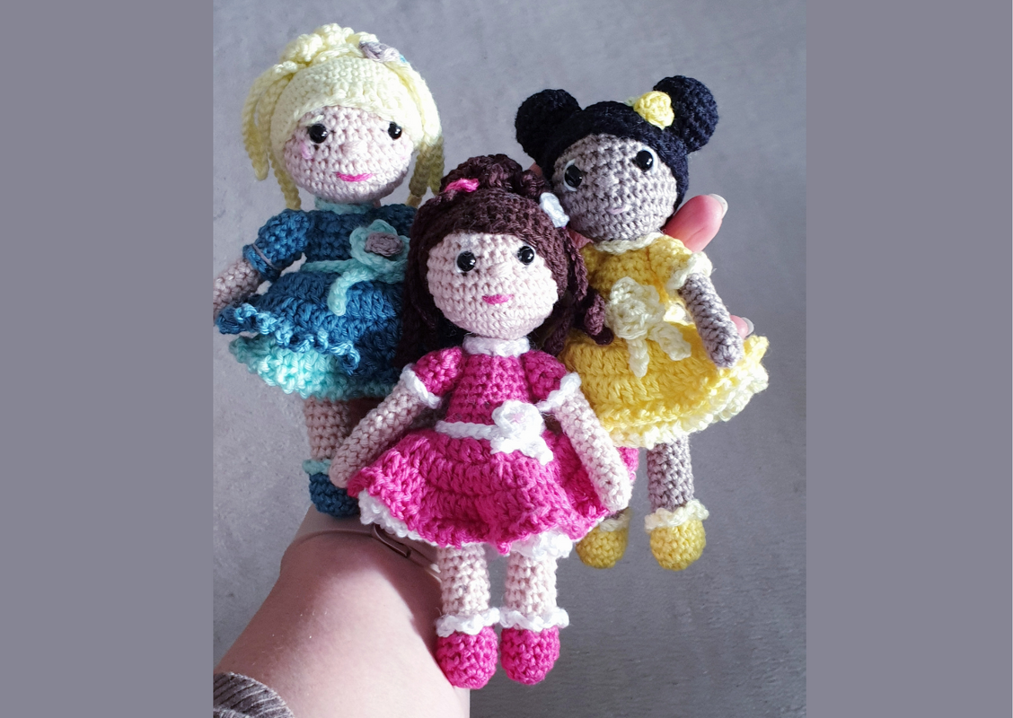Hand hält drei kleine gehäkelte Amigurumi-Puppen in Rosa, Gelb und Blau mit Rüschenkleidern