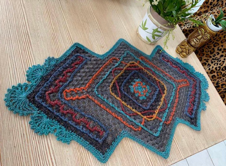 Gehäkelter Tischläufer in Anthrazit mit bunten Rauten- und Zickzackmustern und türkisfarbenem Muschelrand auf einem Holztisch.