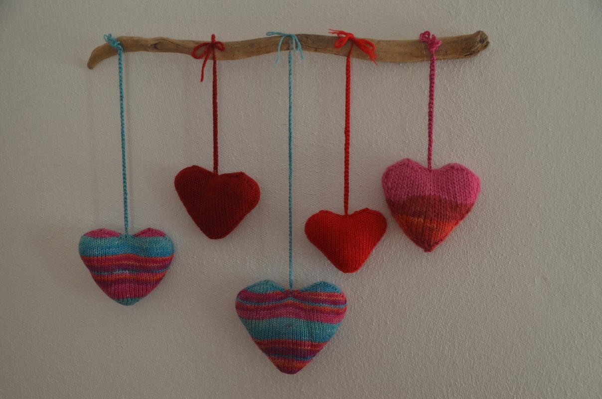 Fünf gestrickte Herzanhänger an einem Treibholzstab, aufgehängt an Schnüren vor einer weißen Wand