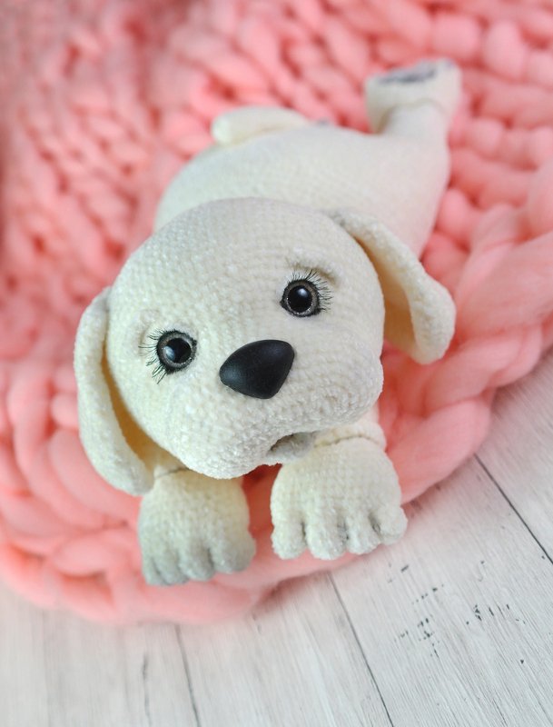 Gehäkelter cremeweißer Amigurumi-Welpe mit schwarzen Augen und Nase auf rosa Decke