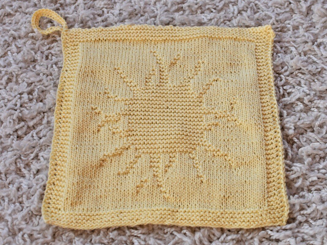 Gelber gestrickter, quadratischer Topflappen mit Sonnenmotiv und Aufhängeschlaufe auf hellem Teppich