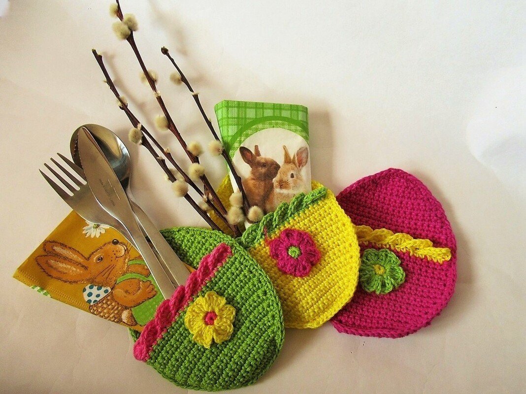 Drei gehäkelte Bestecktaschen in Ei‑Form in Grün, Gelb und Pink mit Blumenapplikationen, neben Besteck, Hasenserviette und Weidenkätzchen.