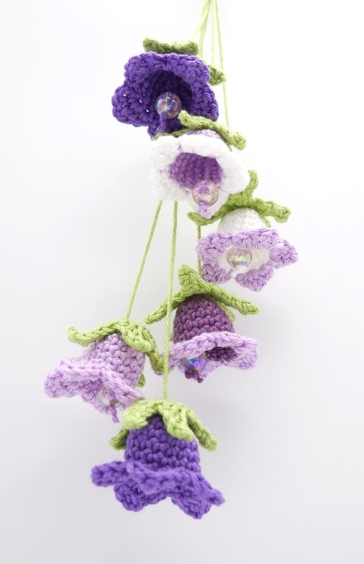 Hängende Kette aus gehäkelten Glockenblumen in Lila, Violett und Weiß mit grünen Kelchblättern und Perlen in der Mitte.