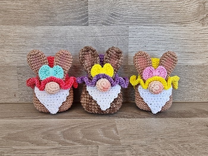 Drei gehäkelte Amigurumi-Osterwichtel mit braunen Hasenohren, weißem Bart, Rüschenkragen und bunten Schleifen vor Holz.