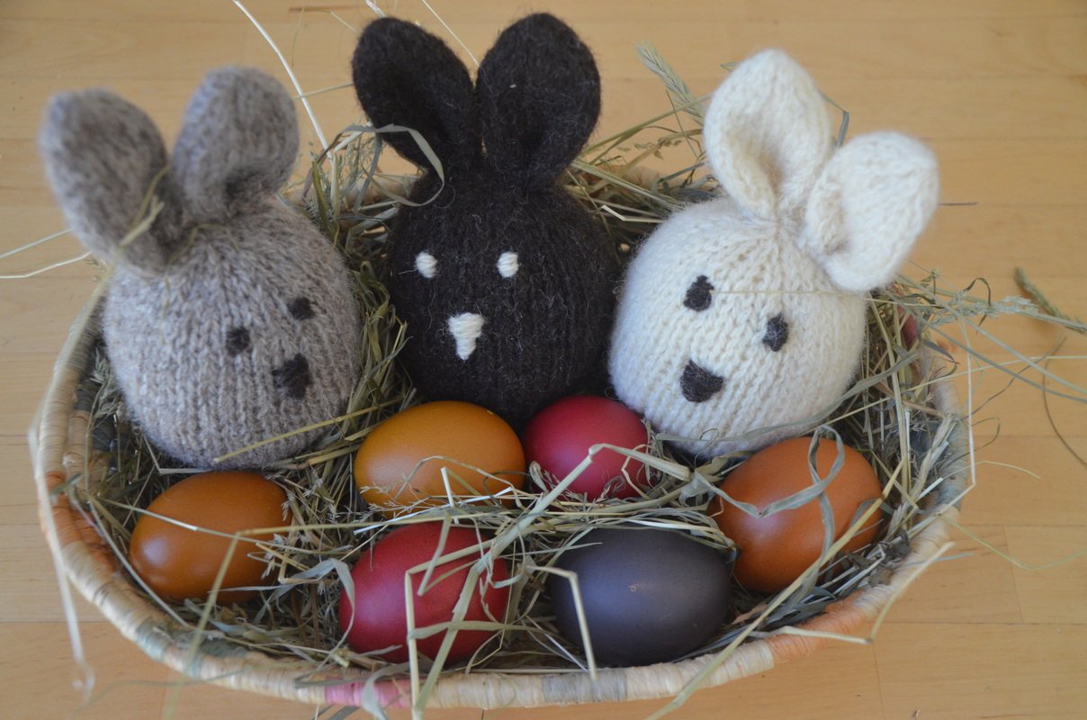 Heukorb mit drei gestrickten Hasen-Eierwärmern (grau, dunkelbraun, weiß) und bunten Ostereiern