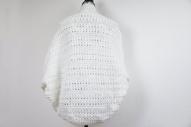 Back view of a white crocheted shrug on a dress form, showing openwork rows and ribbed hem