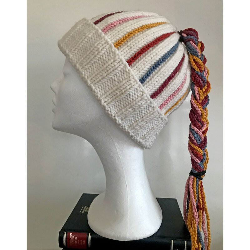 Weiße gestrickte Damenmütze mit bunten Querstreifen und mehrfarbigem, geflochtenem Zopf auf einem Schaufensterkopf.