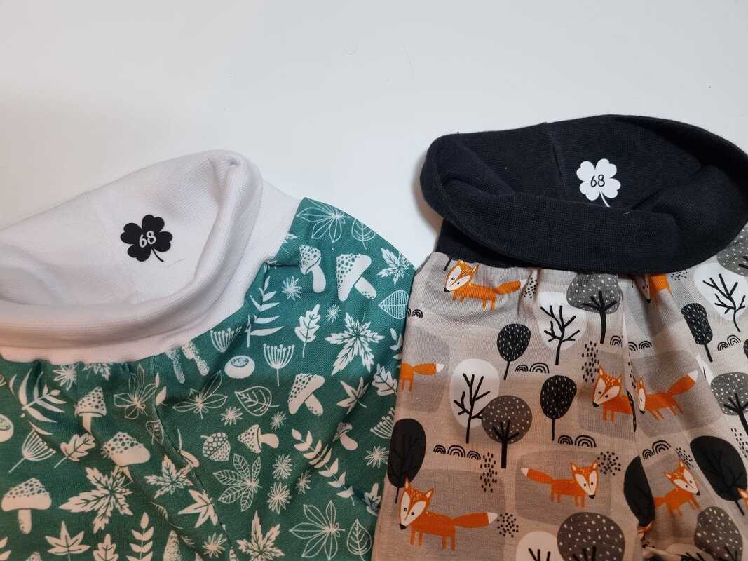 Zwei Kinderhosen mit Bündchen; links grün mit Pilz- und Blattmuster, rechts beige mit orangefarbenen Fuchs- und Baumillustrationen