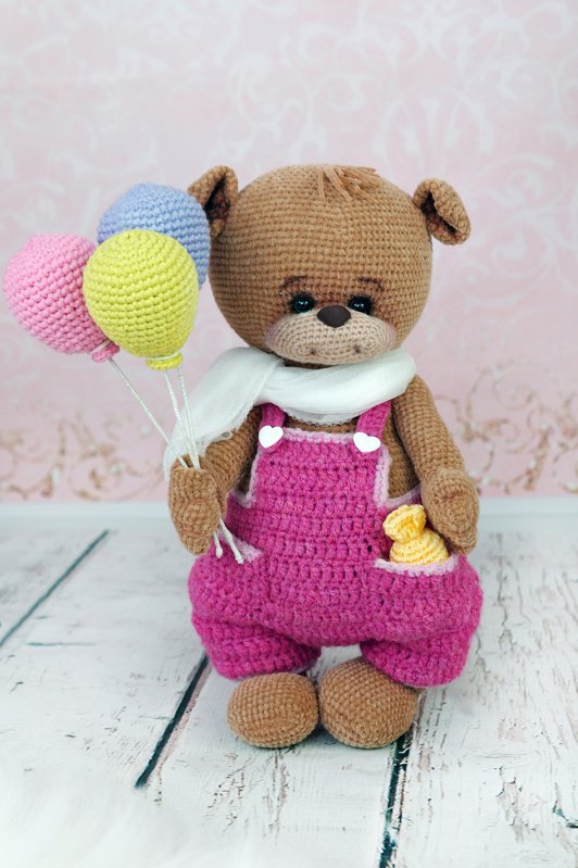 Crocheted brown amigurumi teddy bear wearing pink overalls, white scarf, holding three pastel balloons and a small yellow duck in its pocket.