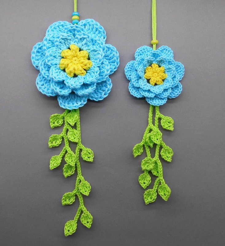 Two crocheted hanging decorations, large and small, each with a blue layered flower, yellow center, and trailing green leaf vines.
