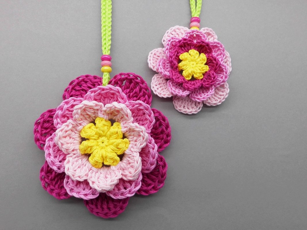 Two layered crochet flower pendants in shades of pink with yellow centers hanging on green cords with beads against a grey background