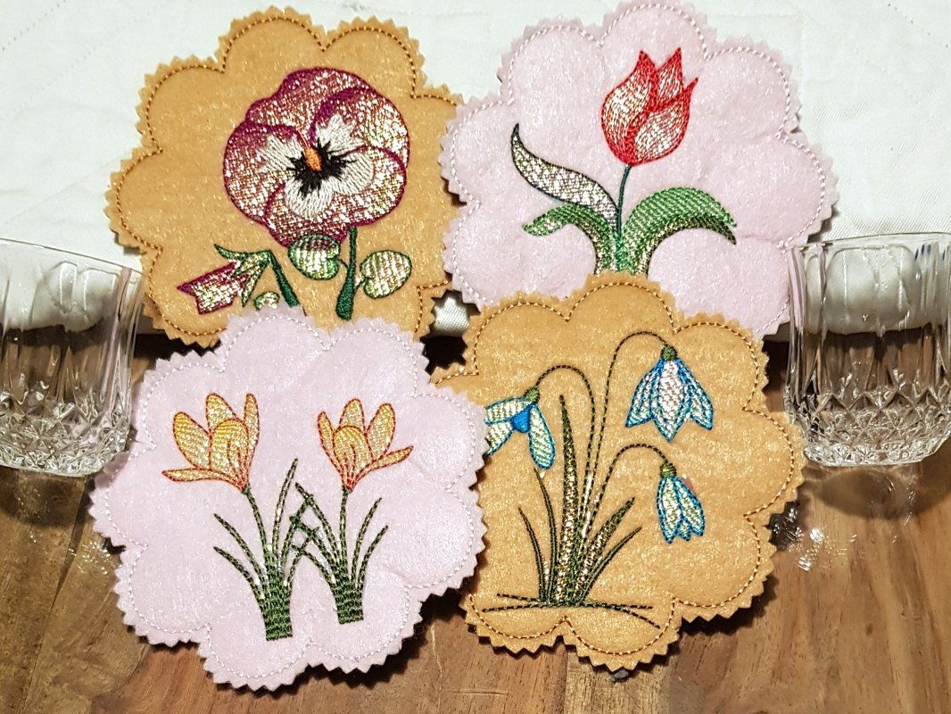 Vier gestickte ITH-Tassenteppiche mit Blumenmotiven in Pastellfarben auf Holzuntergrund