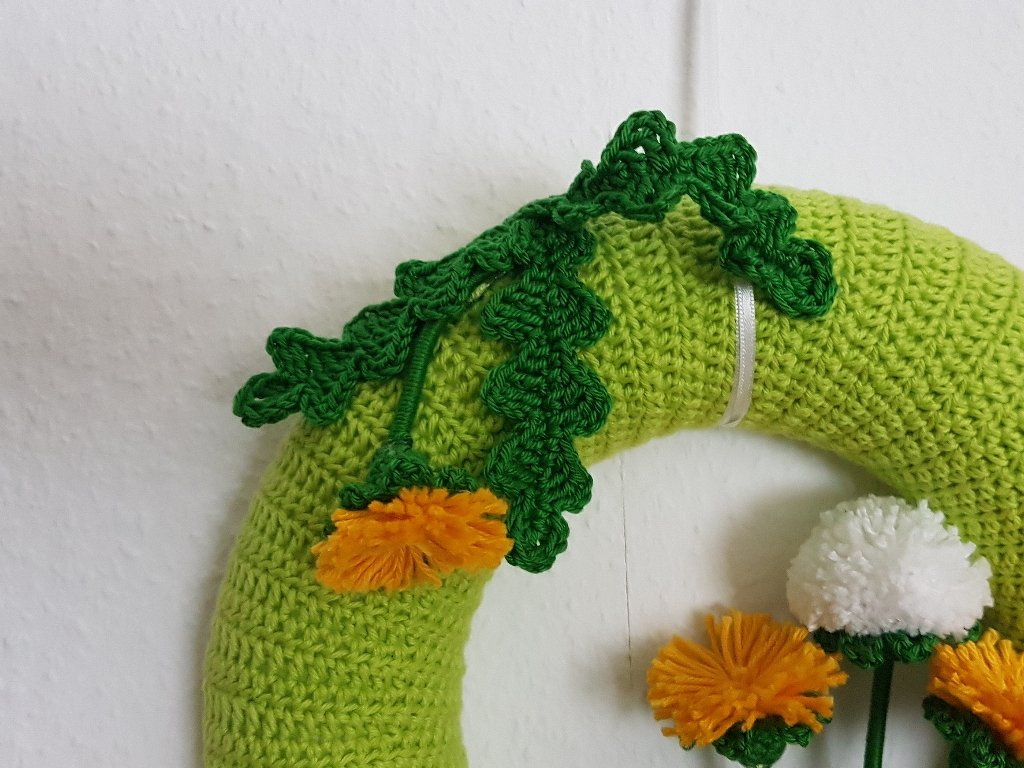 Hellgrüner, handgehäkelter Kranz mit gelben und weißen Löwenzahnblüten und grünen Blättern.