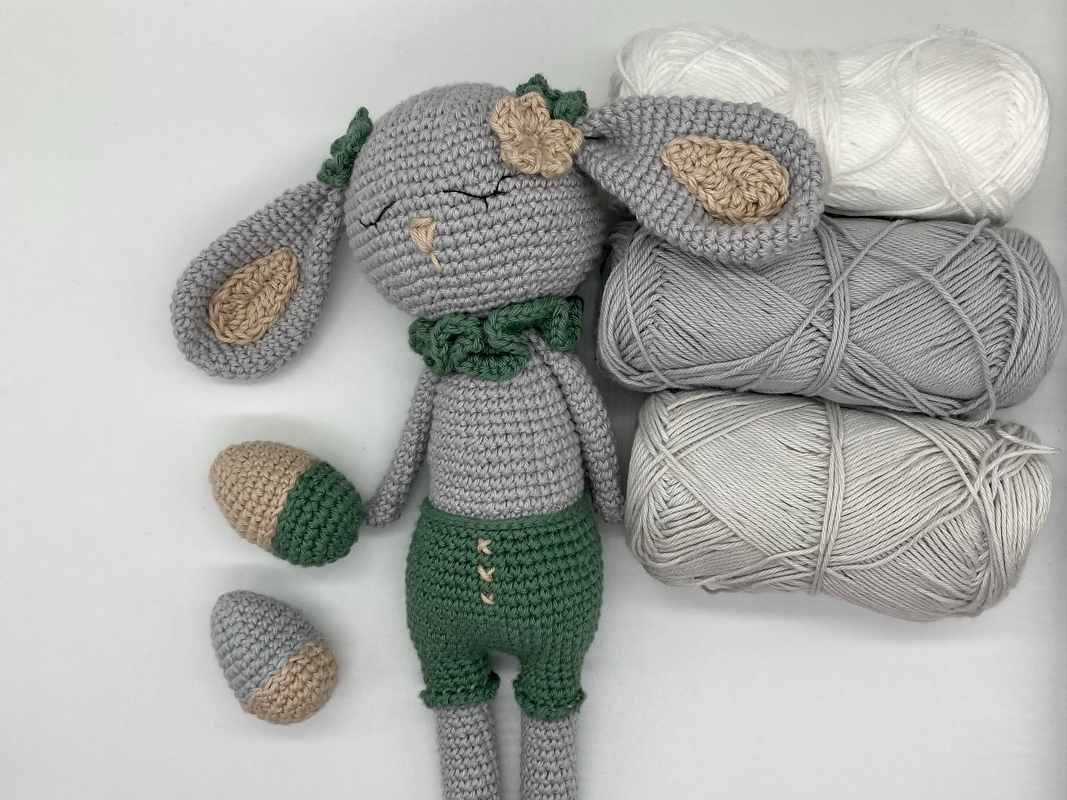 Gehäkelter grauer Amigurumi-Hase mit grüner Hose und zwei gehäkelten Eiern neben Garnknäueln