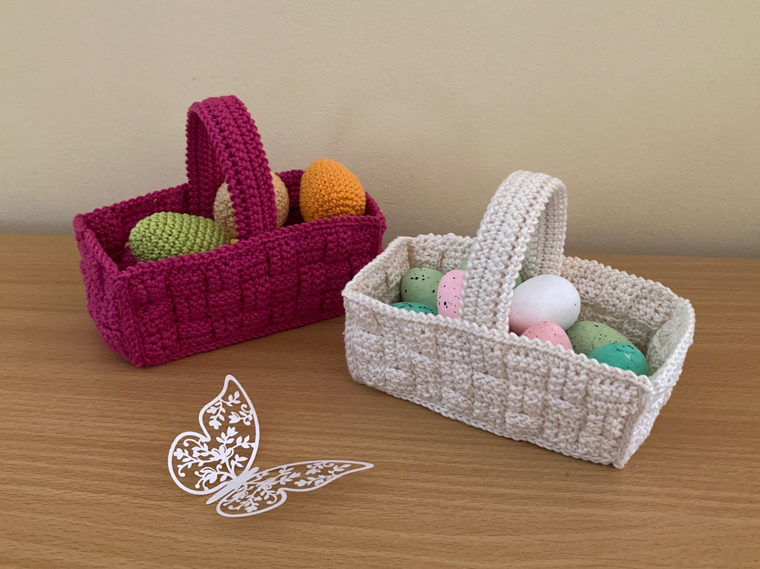 Zwei rechteckige Häkelkörbe mit breitem Henkel, pink und creme, mit bunten Eiern gefüllt auf einem Holztisch; vorne ein weißer Schmetterling.