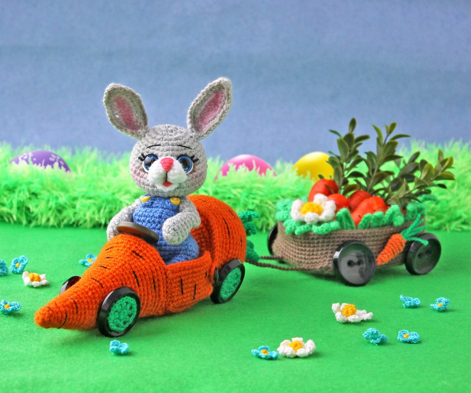 Gray crocheted amigurumi rabbit in blue overalls driving an orange carrot car with button wheels, pulling a small trailer filled with crochet carrots and flowers on a green backdrop.