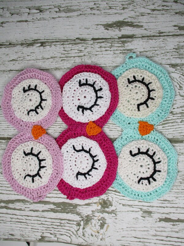 Sechs gehäkelte Eulen-Untersetzer in Rosa, Fuchsia und Türkis auf verwittertem Holz.