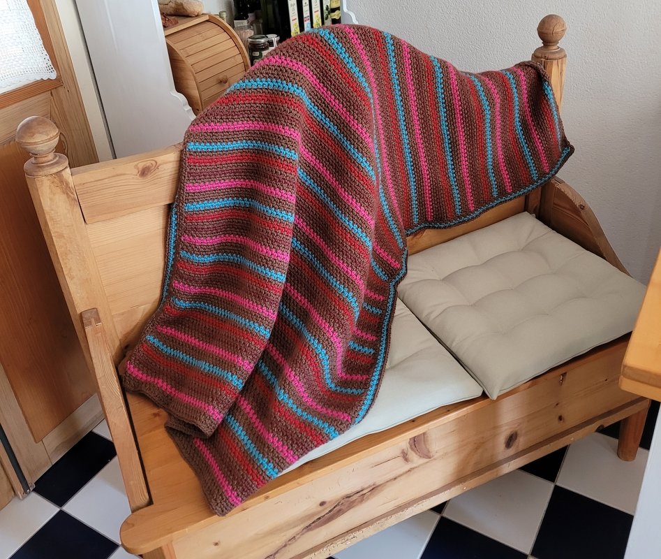 Gehäkelte gestreifte Decke in Braun, Pink und Türkis auf einer hellen Holzbank mit Sitzkissen