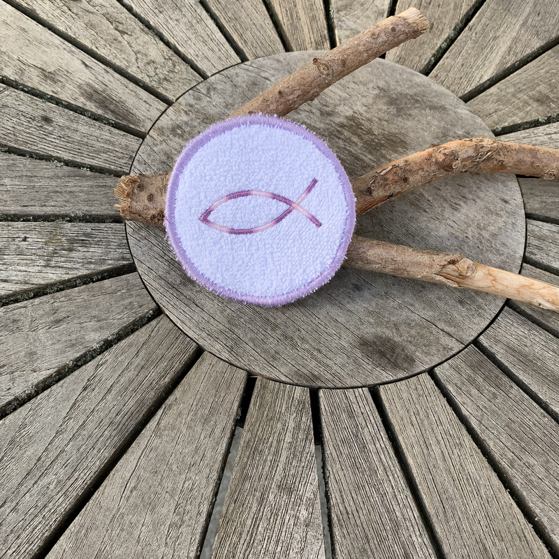 Runder, lilafarbener Untersetzer mit gesticktem Fischsymbol auf hölzerner Oberfläche