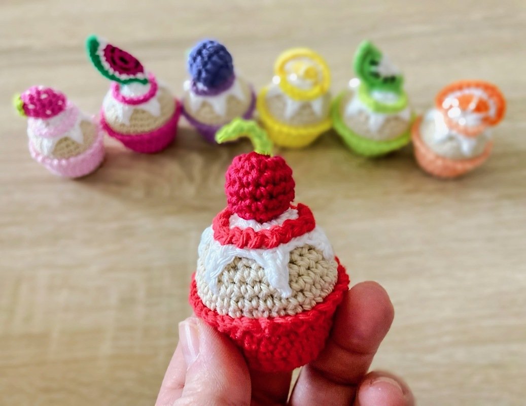 Gehäkelter Cupcake mit roter Kirsche in einer Hand; mehrere gehäkelte Cupcakes mit Fruchtverzierungen im Hintergrund