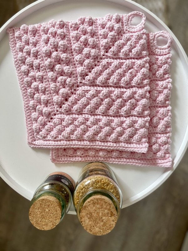 Zwei rosa gehäkelte Topflappen mit Noppenmuster und Aufhängeschlaufen auf einer weißen Ablage, daneben Gewürzgläser mit Korkdeckeln.
