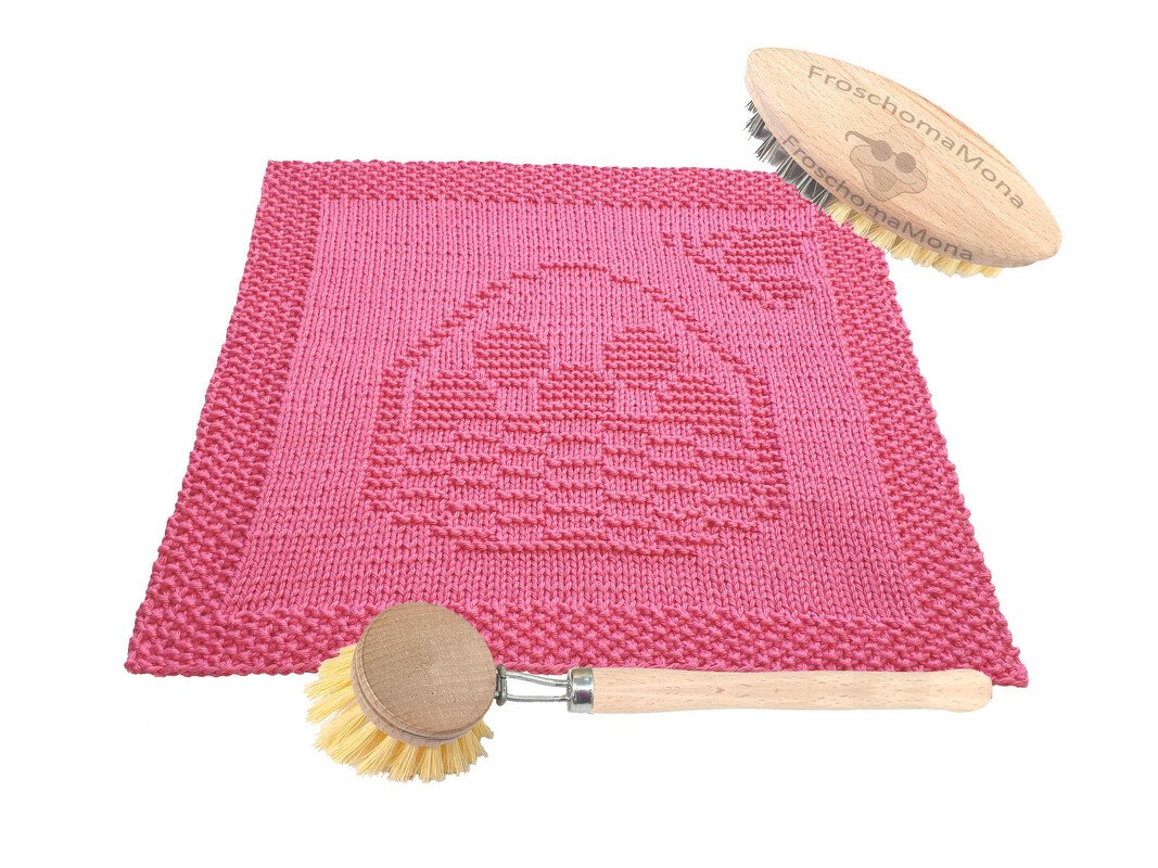 Rosa gestrickter quadratischer Topflappen mit Hasenmotiv, daneben zwei Holzspülbürsten auf hellem Hintergrund.