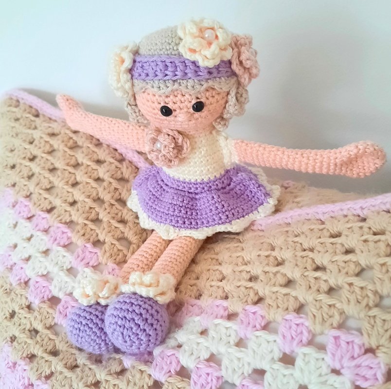 Gehäkelte Amigurumi-Puppe mit lila Rock, cremefarbenem Oberteil und Blumen im Haar auf beigefarben‑rosa gestreifter Häkeldecke.