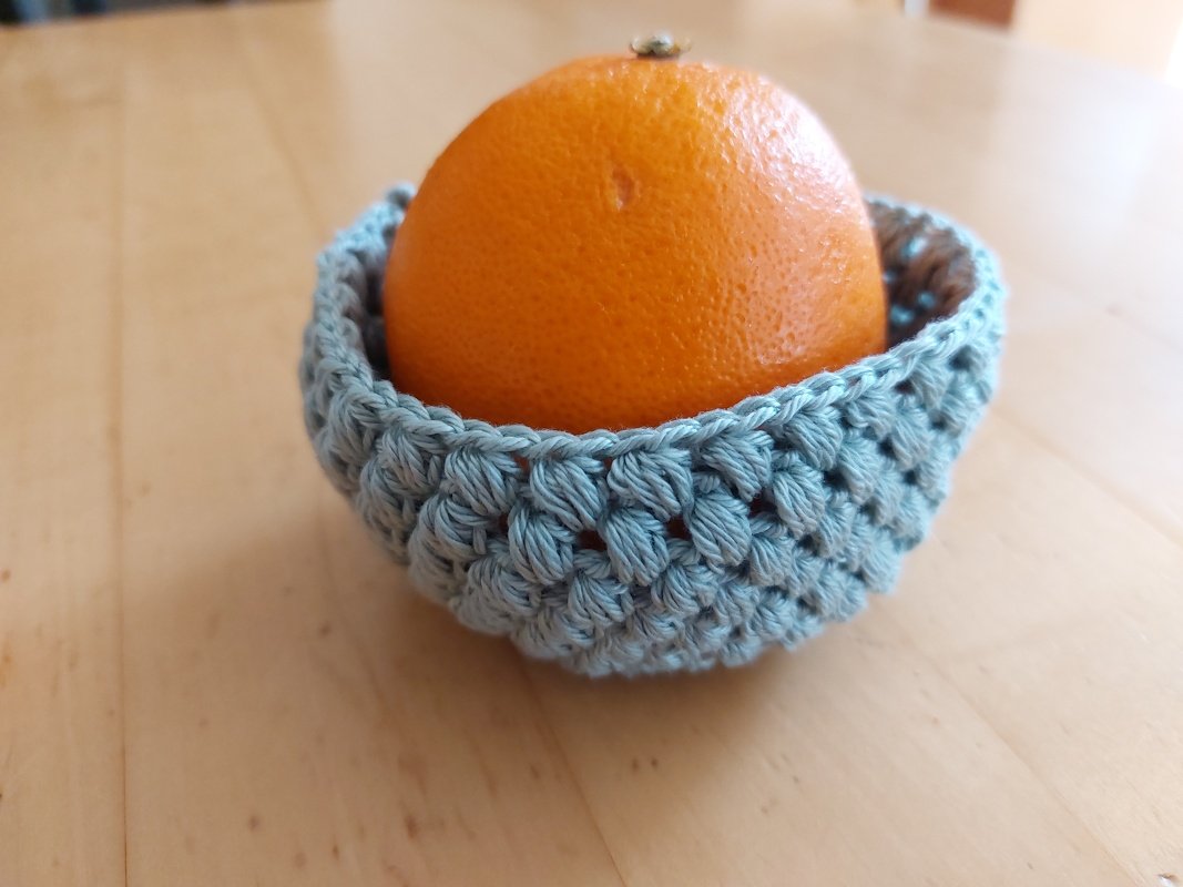 Orange in einem kleinen helltürkisen gehäkelten Korb mit Puffmaschen auf einem Holztisch.