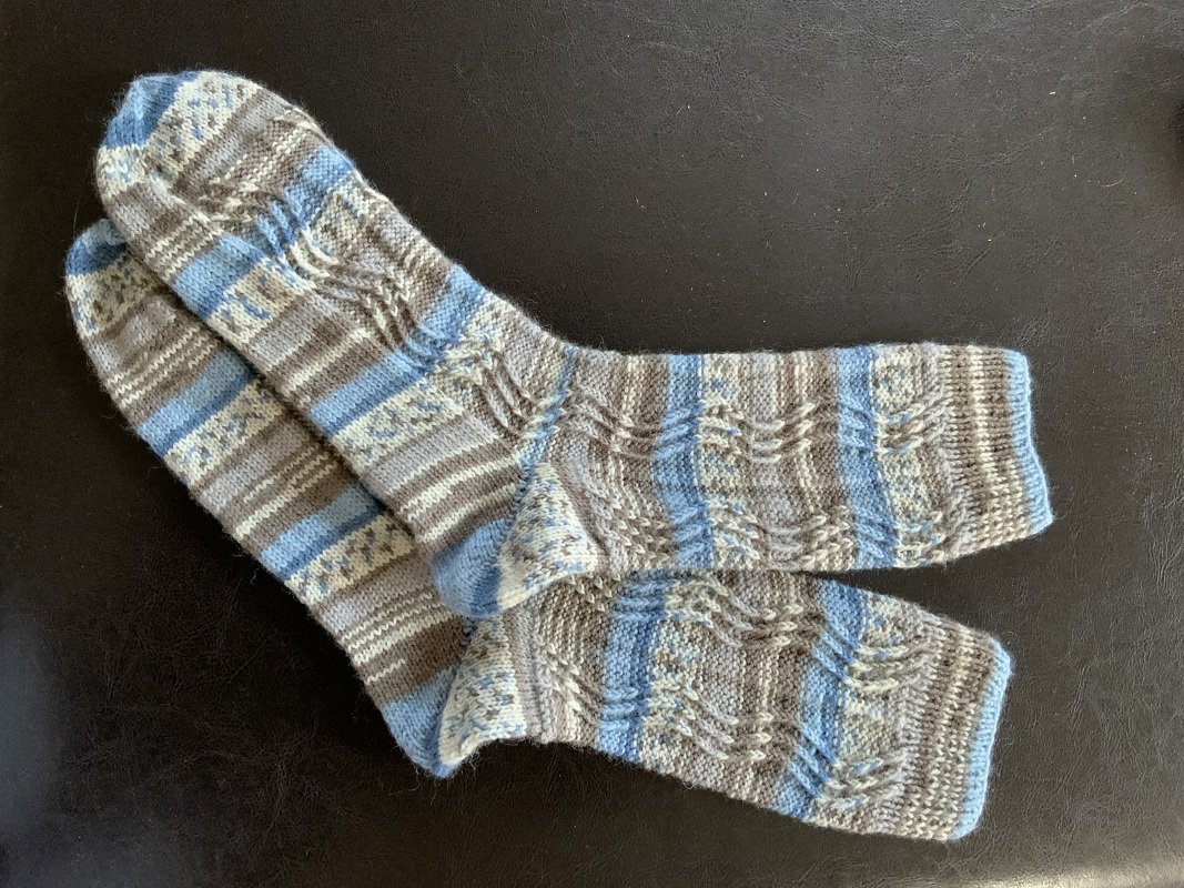Handgestricktes Paar Socken in Blau-, Grau- und Hellgrautönen mit Streifen- und Strukturmuster auf dunklem Hintergrund.