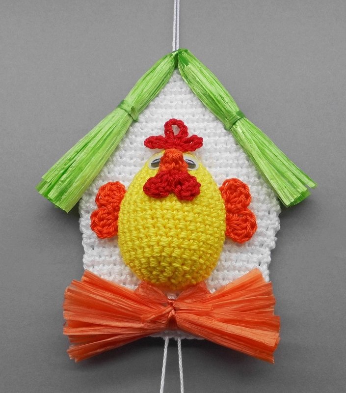 Yellow crocheted chick with red comb and wattle, on a white house-shaped crochet backing, with green raffia roof and orange raffia bow, and a hanging loop.